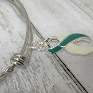 May include: A silver bangle bracelet with a teal and white ribbon charm. The bracelet is made of twisted metal and has a clasp closure.