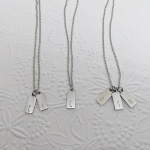 May include: Three silver necklaces with personalized initial tags. The first necklace has two tags with the letters "C" and "V". The second necklace has one tag with the letter "C". The third necklace has three tags with the letters "O", "S", and "M".