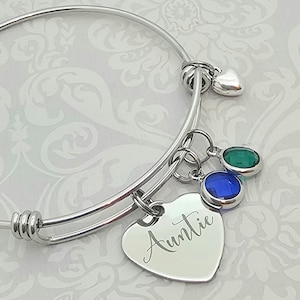 May include: A silver bangle bracelet with a heart charm engraved with the word "Auntie". The bracelet has two dangling charms with blue and green gemstones.