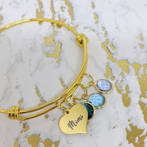 May include: Gold-toned bangle bracelet with a heart-shaped charm engraved with "Mimi". The bracelet features three gemstone charms in shades of blue. The bracelet is displayed on a white and gold marble-patterned surface.
