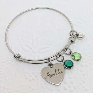 Op de afbeelding: Een zilveren armband met een hartvormige bedel met de tekst "Abuelita" en drie groene kristallen bedels.