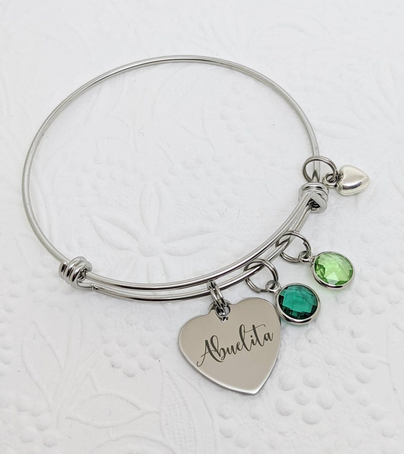 Op de afbeelding: Een zilveren armband met een hartvormige bedel met de tekst "Abuelita" en drie groene kristallen bedels.