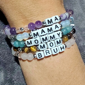 May include: A stack of bracelets with white letter beads spelling out "MA", "MAMA", "MOMMY", "MOM", and "BRUH". The bracelets are made with amethyst, agate, tiger's eye, and other beads.