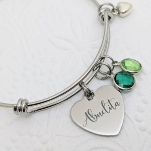 Op de afbeelding: Een zilveren uitbreidbare armband met een hartbedel gegraveerd met het woord "Abuelita". De armband heeft ook twee groene kristallen bedels en een kleine zilveren hartbedel.