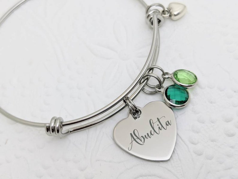 Op de afbeelding: Een zilveren uitbreidbare armband met een hartbedel gegraveerd met het woord "Abuelita". De armband heeft ook twee groene kristallen bedels en een kleine zilveren hartbedel.