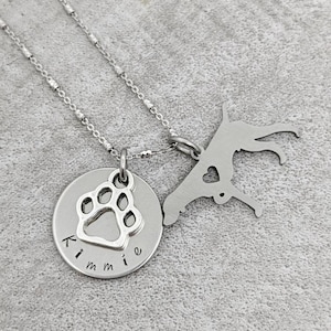 May include: A silver necklace with two pendants. One pendant is a round disc with a paw print and the name "Kimmie". The other pendant is a dog silhouette with a heart cutout. The necklace chain is delicate.