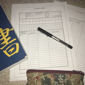 May include: A black and white journal with the title "My Kanji Journal" printed on the cover. The journal has a grid layout with columns for key word, stroke number, radical, memory trick, pronunciation, example sentence, and kanji. A black pen is lying on top of the journal.