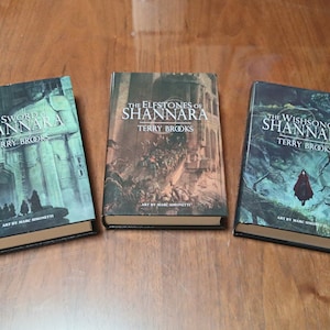 Puede incluir: Tres libros de tapa dura con portadas de arte fantástico. Los títulos incluyen "The Sword of Shannara", "The Elfstones of Shannara" y "The Wishsong of Shannara" de Terry Brooks. Las portadas presentan ilustraciones de Marc Simonetti.