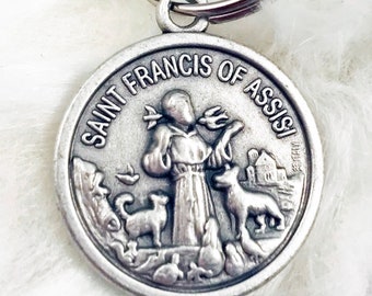 May include: Silver-toned keychain featuring a round medal with a depiction of Saint Francis of Assisi. The medal has the words "Saint Francis of Assisi" inscribed around the edge. The saint is depicted holding a bird in his hand, with other animals surrounding him.
