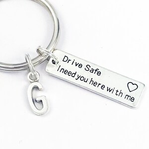 May include: Silver keychain with a charm that says "Drive Safe I need you here with me" and a heart. The charm is attached to a silver key ring with a letter G charm.