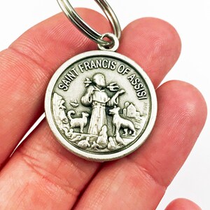 May include: Silver keychain featuring a round medal with a depiction of Saint Francis of Assisi. The medal has the words "Saint Francis of Assisi" inscribed around the edge. The saint is depicted holding a bird and surrounded by animals.