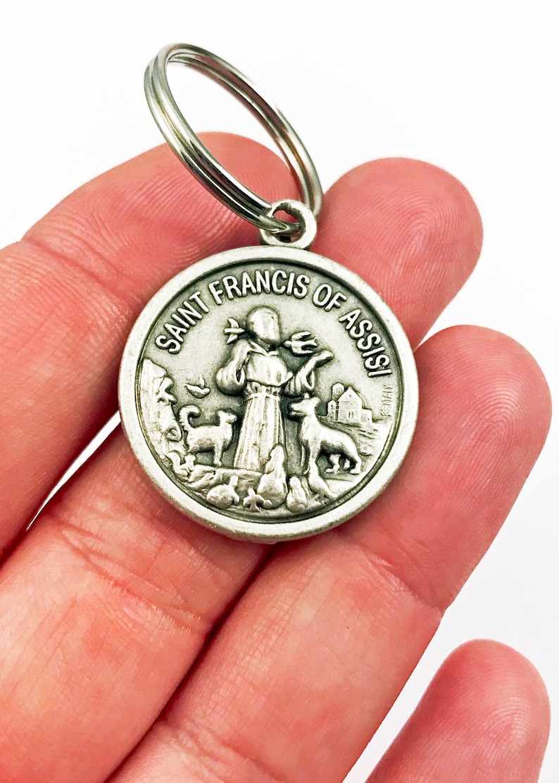 May include: Silver keychain featuring a round medal with a depiction of Saint Francis of Assisi. The medal has the words "Saint Francis of Assisi" inscribed around the edge. The saint is depicted holding a bird and surrounded by animals.