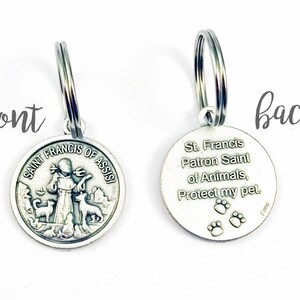 May include: Silver key chain with a round charm featuring Saint Francis of Assisi holding a bird and two dogs. The back of the charm reads "St. Francis Patron Saint of Animals, Protect my pet." with three paw prints.