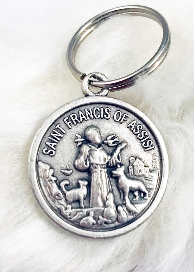 May include: Silver-toned keychain featuring a round medal with a depiction of Saint Francis of Assisi. The medal has the words "Saint Francis of Assisi" inscribed around the edge. The saint is depicted holding a bird in his hand, with other animals surrounding him.