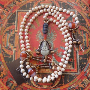 May include: White bead mala necklace with a carved stone pendant featuring a seated Buddha figure. The pendant is surrounded by smaller beads and metal accents. The necklace is displayed on a red and gold mandala background.