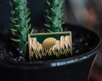 May include: Gold-toned enamel pin with a green and beige mountain scene. The pin features a stylized sun and a geometric pattern of trees.