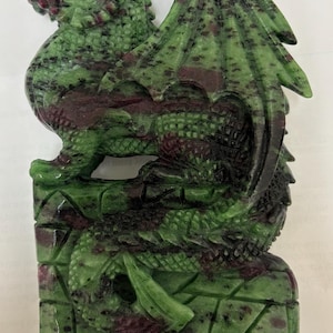 May include: A carved stone dragon sculpture in shades of green and burgundy. The dragon has detailed wings and is perched on a rectangular base. The sculpture is made of a speckled stone material.
