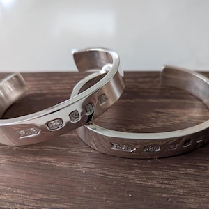 Peut inclure: Deux bracelets de manchette en argent avec poinçons, posés sur une surface en bois. Les bracelets ouverts ont une finition polie. Les poinçons incluent des poinçons de fabricant et un poinçon 925, indiquant de l'argent sterling. Diamètre d'environ 6 cm.