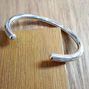 May include: A silver cuff bracelet with a polished finish and rounded terminals. The open design allows for easy wear. The bracelet is displayed on a wooden surface, highlighting its reflective qualities. A simple, elegant piece of jewellery.