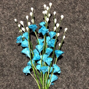 Può includere: Un bouquet di fiori di carta blu con steli e boccioli bianchi. I fiori sono disposti a grappolo e hanno un aspetto delicato e fantasioso.