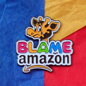 Pode incluir: Um broche de esmalte colorido com uma girafa de desenho animado com uma lágrima, as palavras "BLAME amazon" e uma estrela.