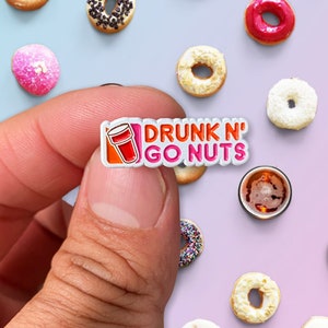 Könnte beinhalten: Eine weiße Emaille-Anstecknadel mit einem rosafarbenen und orangen Donut-Becher und dem Text "DRUNK N' GO NUTS".