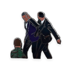 Puede incluir: Un pin de esmalte en blanco y negro que representa a dos hombres con traje, uno de los cuales está extendiendo la mano para chocar los cinco con el otro. El hombre del fondo lleva una camisa verde.