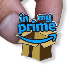 May include: A silver pin with a blue and white Amazon Prime logo and the text "in my prime" on a brown cardboard box.