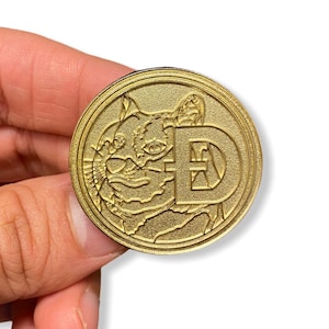 May include: A gold-colored metal coin with a detailed image of a cat's face and the letter "E" in the center.
