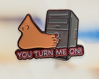May include: A red and black enamel pin featuring a cartoon foot pressing a red button on a gray computer tower. The text "YOU TURN ME ON!" is written in white on a red background.