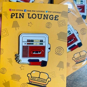 Może przedstawiać: Emaliowana przypinka przedstawiająca biały i czerwony odtwarzacz kasetowy z mikrofonem i żółtą kanapą z napisem "PIN LOUNGE". Tło jest żółte z różnymi bazgrołami.