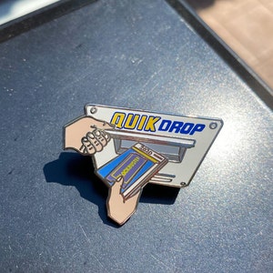 Könnte beinhalten: Ein silberfarbener Emaille-Pin mit einem "QUIK DROP"-Schild, einer Hand, die eine Blockbuster-DVD in einen Schlitz steckt, und einer anderen Hand, die die DVD hält. Die DVD-Hülle ist blau und gelb, mit dem Jahr 2020.
