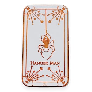 May include: A white enamel pin with a copper-colored outline. The pin features a tarot card design with the Hanged Man card. The card depicts a stylized figure hanging upside down with a halo and a spider web. The card is surrounded by a border of stars.