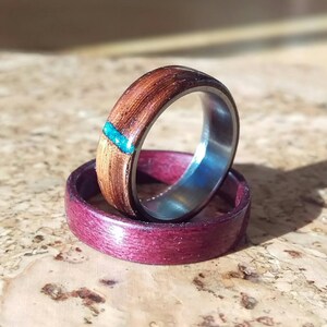 May include: Two wooden rings with a turquoise inlay. The top ring is brown wood with a turquoise inlay. The bottom ring is a deep purple wood.
