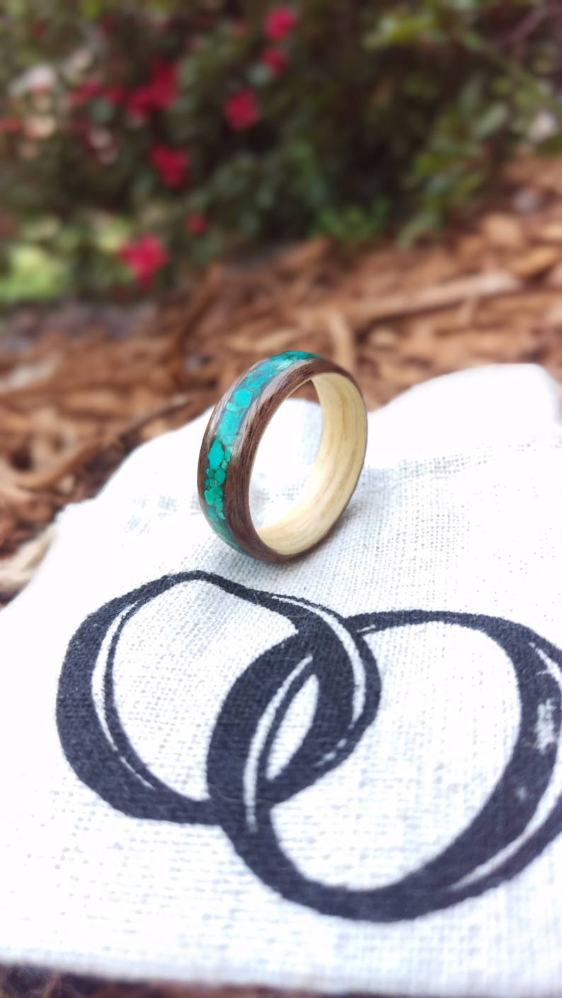 May include: A wooden ring with a turquoise inlay. The ring is on a white fabric background with a black design of two interlocking circles.