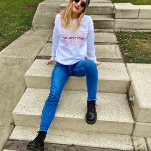 May include: A white sweatshirt with the text "CARE ABOUT OTHERS" repeated in a stacked pattern. The model is wearing blue jeans and black boots.