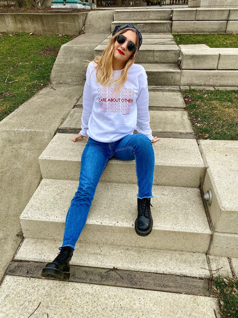 May include: A white sweatshirt with the text "CARE ABOUT OTHERS" repeated in a stacked pattern. The model is wearing blue jeans and black boots.