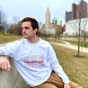 May include: White sweatshirt with the phrase "CARE ABOUT OTHERS" repeated in a gradient pattern from red to white. The text is stacked vertically and the phrase "Have a Nice Day" is at the bottom.