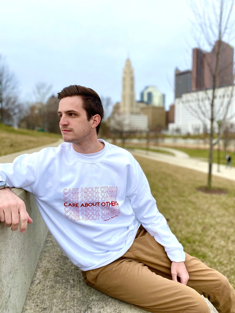 May include: White sweatshirt with the phrase "CARE ABOUT OTHERS" repeated in a gradient pattern from red to white. The text is stacked vertically and the phrase "Have a Nice Day" is at the bottom.
