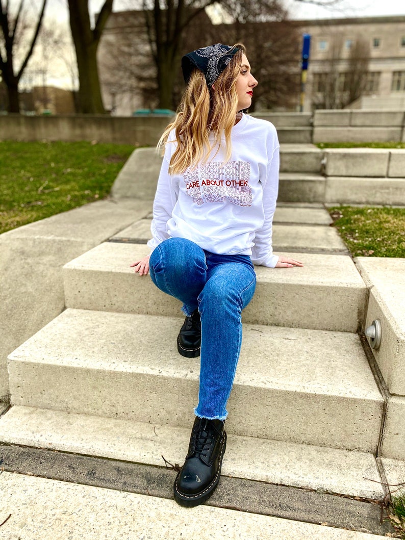 May include: A white long-sleeve sweatshirt with the text "CARE ABOUT OTHERS" repeated in a stacked pattern. The sweatshirt is worn by a person sitting on a set of concrete stairs. The person is wearing blue jeans and black boots.