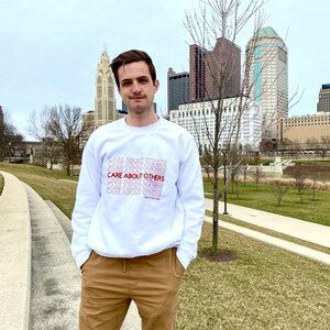 May include: A white sweatshirt with the phrase "CARE ABOUT OTHERS" repeated in red and pink lettering. The person wearing the sweatshirt is standing in front of a city skyline.