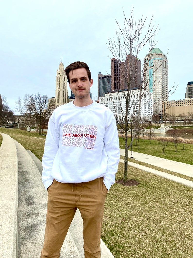 May include: A white sweatshirt with the phrase "CARE ABOUT OTHERS" repeated in red and pink lettering. The person wearing the sweatshirt is standing in front of a city skyline.