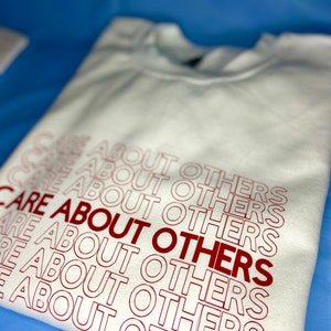 May include: White sweatshirt with the phrase "CARE ABOUT OTHERS" repeated in red lettering.