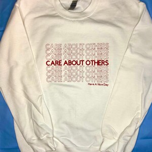 May include: A white sweatshirt with the phrase "CARE ABOUT OTHERS" repeated multiple times in red lettering. The text "Have A Nice Day" is printed in smaller red lettering below the repeated phrase.