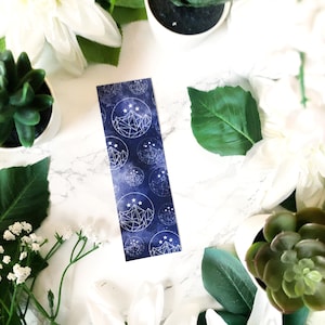 May include: A blue bookmark featuring a white geometric mountain design and stars. The bookmark is surrounded by white flowers and green succulents.