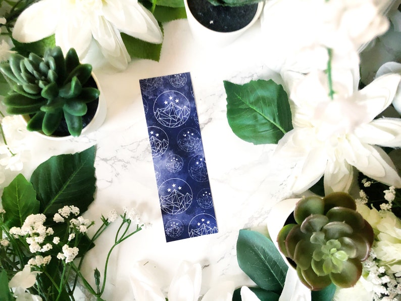 May include: A blue bookmark featuring a white geometric mountain design and stars. The bookmark is surrounded by white flowers and green succulents.