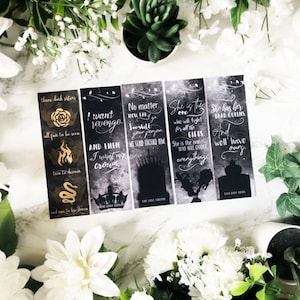 May include: Set of five black and gold bookmarks with quotes from the book "Six of Crows" by Leigh Bardugo. The quotes are: "These dark sisters all fear to be seen", "I want revenge", "And then I want my crown", "No matter how far I fall I will stand beside you", "She is the one who will fight for all the gifts. She is the one who will change everything", "She has her dead queens. And well have ours."