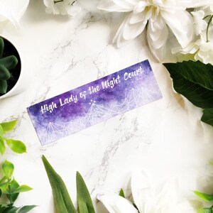 May include: A purple and white bookmark with the text "High Lady of the Night Court" printed on it. The bookmark features a watercolour background with a starry sky and mountains.