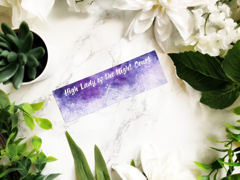 May include: A purple and white bookmark with the text "High Lady of the Night Court" printed on it. The bookmark features a watercolour background with a starry sky and mountains.