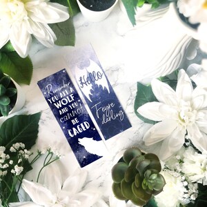 May include: Two blue and white bookmarks with a galaxy background. One bookmark features a wolf silhouette and the text "Remember you are a wolf and you cannot be caged". The other bookmark features a bat silhouette and the text "Hello Feyre darling".
